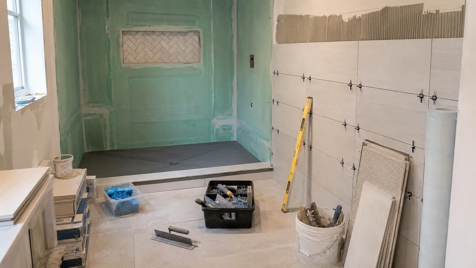 Bathroom mid-remodel at the waterproofing stage, with green membrane sealing the shower walls, a sloped shower pan set, and large-format wall tile going up with spacers and a level